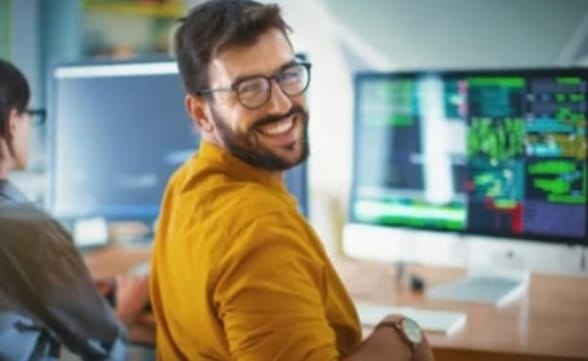 A guy with short brown hair and a beard, wearing glasses and a mustard-yellow shirt, is turned towards the camera with a smile. He's in an office with computers on a desk, and another person is partially visible on the left.