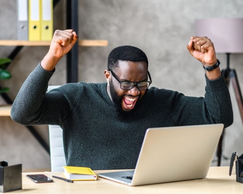 A man with short black hair and a beard, wearing a dark green sweater and black glasses, sits at a desk with a laptop. He's raising both fists in celebration with his mouth open in excitement. The desk has a phone, notebook, pen, and other items. A lamp and bookshelves are in the background.