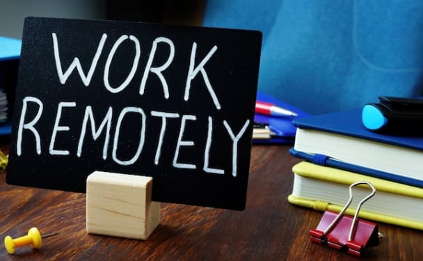A black chalkboard sign with "WORK REMOTELY" written in white chalk on a wooden desk. The sign is propped up by a small wooden cube. There's a stack of books (blue, yellow) with a red binder clip on the right, a yellow pushpin on the left, and pens in the background against a blue wall.