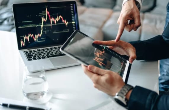 Looks like some folks are crunchin' numbers and checkin' out market trends! 📈 They're got a laptop showin' a graph with red and green lines - probably stock prices or somethin'. One person's pointin' at a tablet with a similar graph. Guess they're discussin' investments or business stuff. What do you think about this scene?