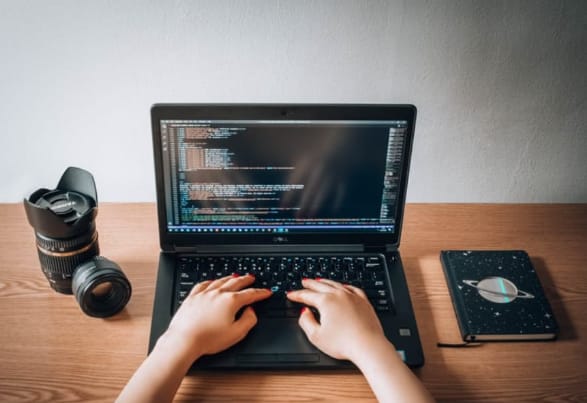 Alt text: A person typing on a laptop with code on the screen, sitting at a wooden table with a camera lens and a notebook with a planet design beside the laptop.