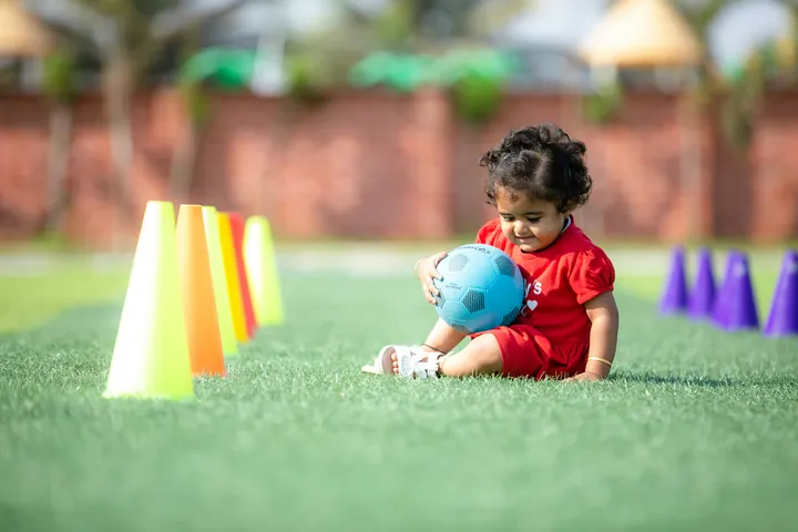 A young child in a red dress sitting on grass with a football, with colorful boulders on each side.
